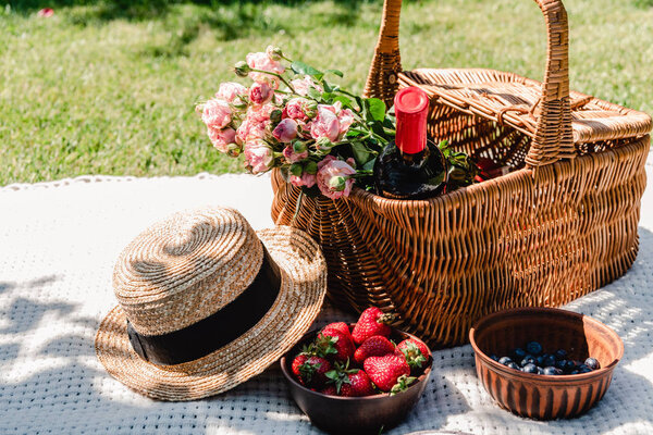 wicker basket with roses and bottle of wine on white blanket near straw hat and berries at sunny day in garden