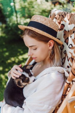 bahçede şezlongda otururken köpek yavrusu tutan hasır şapka sarışın kız