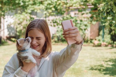 yeşil bahçede sarışın kız kapalı gözleri ve sevimli köpek yavrusu ile selfie alarak