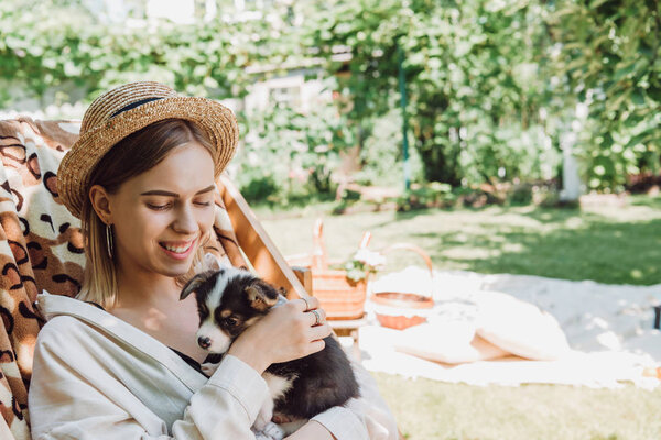 smiling blonde girl in straw hat holding puppy while sitting in deck chair in green garden