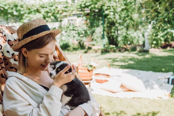 yeşil bahçede şezlongda otururken köpek yavrusu tutan hasır şapka mutlu sarışın kız