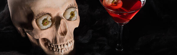 panoramic shot of red cocktail near spooky skull on black background