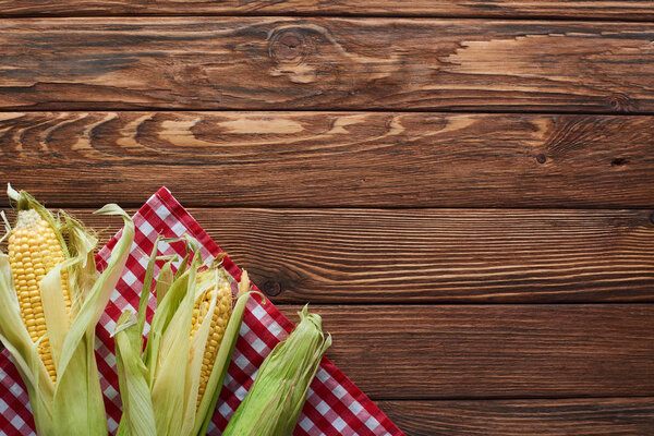 вид сверху на сырую кукурузу на клетчатой скатерти на деревянной поверхности
