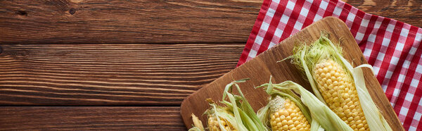 panoramic shot of cutting board with raw corn on checkered tablecloth on wooden surface