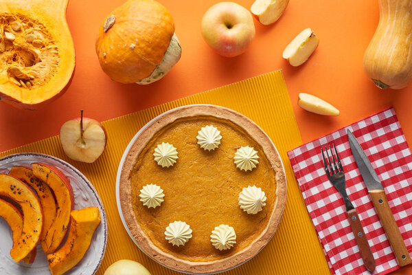 tasty pumpkin pie with whipped cream near baked and raw pumpkins, whole and cut apples, fork and knife on orange surface