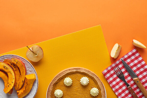 delicious pumpkin pie with whipped cream near baked pumpkin, cut apple, fork and knife on orange surface