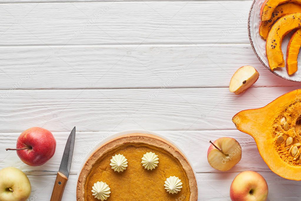 tasty pumpkin pie with whipped cream near baked pumpkin, whole and cut apples, and knife on white wooden surface