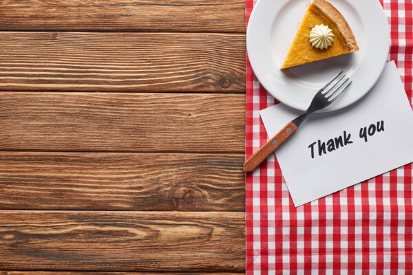 top view of piece of pumpkin pie on plate with fork and thank you card on wooden brown table with red plaid napkin