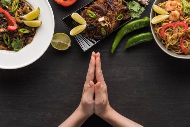 Tahta gri yüzeyde baharatlı Tayland eriştesinin yanında dua eden ellerin kesilip biçilmiş görüntüsü.