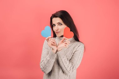 Pembe üzerine mavi ve kırmızı kalp şeklinde kağıt çizimler tutan kazaklı çekici bir kadın. 
