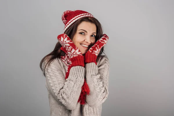 cheerful woman in gloves and knitted hat touching face isolated on grey