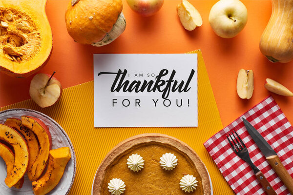 top view of pumpkin pie, ripe apples and card with i am so thankful for you illustration on orange background