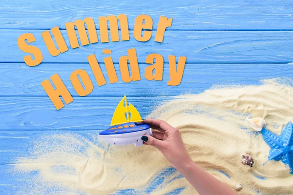 Manos femeninas con barco de juguete por inscripción de vacaciones de verano sobre fondo de madera azul - foto de stock