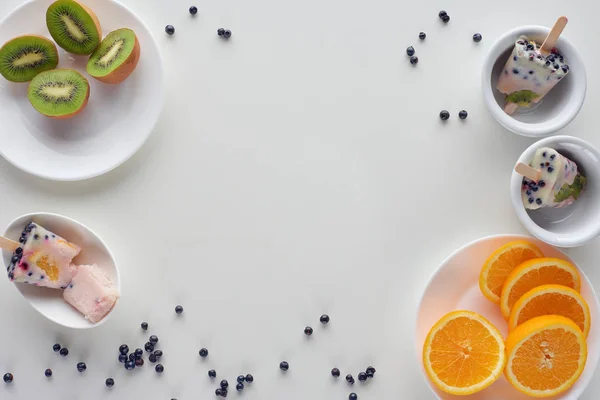 Vista superior de saborosos picolés caseiros em tigelas, fatias de laranja e kiwi em pratos e bagas em fundo cinza — Fotografia de Stock