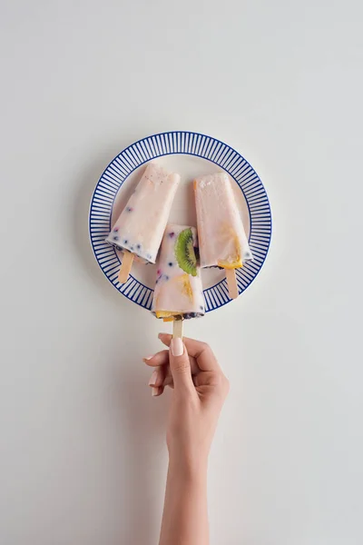 Cropped shot of person holding delicious fruity popsicle above plate on grey — Stock Photo