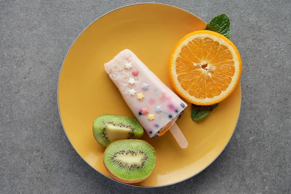 Draufsicht auf köstliches Eis am Stiel mit Orangen- und Kiwi-Scheiben auf gelbem Teller auf grau — Stockfoto