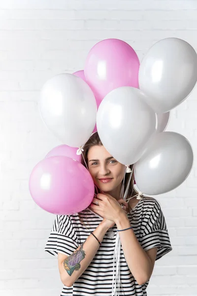 Donna sorridente che tiene palloncini rosa e bianchi su sfondo muro di mattoni bianchi — Foto stock