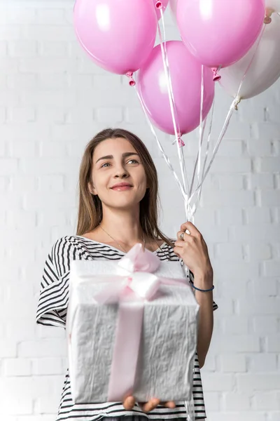 Mujer sonriente sosteniendo caja de regalo y globos sobre fondo de pared de ladrillo blanco - foto de stock