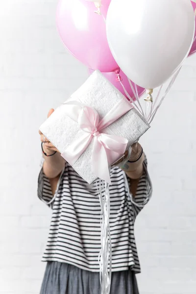 Woman holding gift box and balloons in front of her face on white brick wall background — Stock Photo