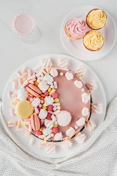 Acostado plano con pastel de cumpleaños rosa, malvaviscos, cupcakes y batidos en la mesa con mantel - foto de stock