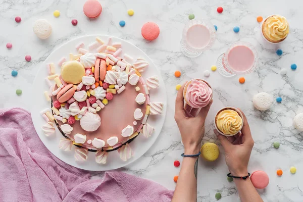 Vista cortada de mãos com cupcakes na mesa com bolo de aniversário rosa e batidos de leite em óculos — Fotografia de Stock