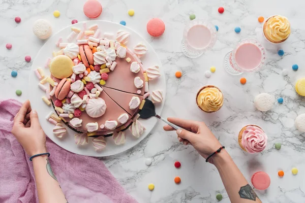 Cropped view of woman taking piece of pink birthday cake with marshmallows and macarons — Stock Photo