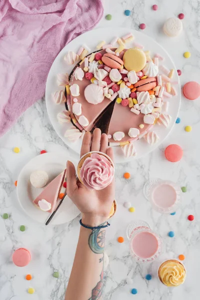 Cropped view of tattooed hands with cupcake over marble table with pink birthday cake, candies, marshmallows and milkshakes — Stock Photo