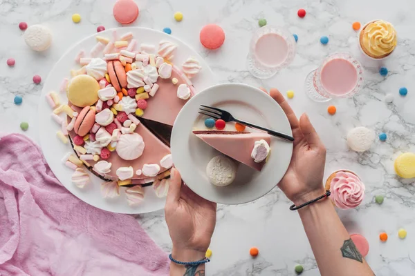 Cropped view of woman holding fork and plate with piece of pink birthday cake and macaron — Stock Photo