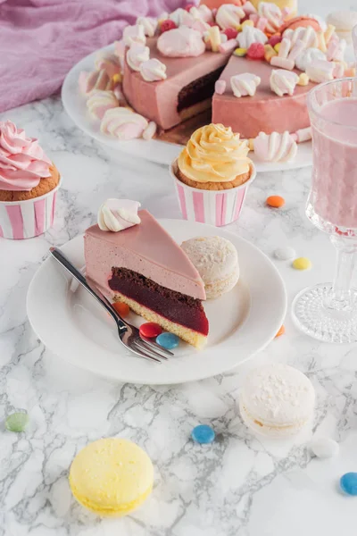 Pedazo de pastel de cumpleaños, macarrones, cupcakes de colores y batido en vidrio en la mesa - foto de stock