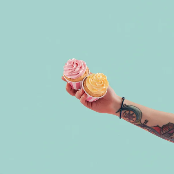 Cropped view of tattooed girl holding two cupcakes with buttercream isolated on blue — Stock Photo