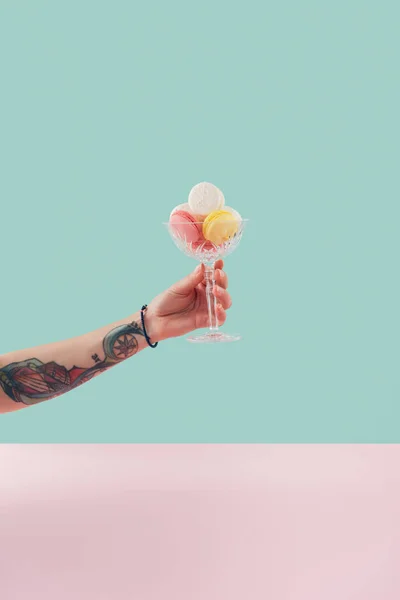 Cropped view of tattooed hand with macarons in glass bowl on pastel background — Stock Photo