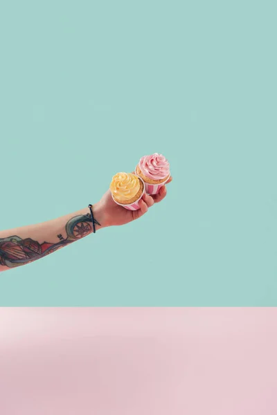 Vista recortada de mujer tatuada sosteniendo dos cupcakes con crema de mantequilla - foto de stock