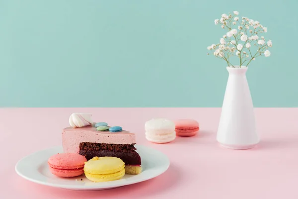 Macaroons and one piece of cake on plate with flowers in vase — Stock Photo