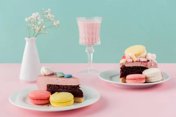 Macaroons and pieces of cake on plates with milkshake in glass and flowers in vase — Stock Photo