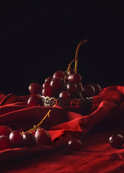 Close-up shot of ripe grapes in vintage metal bowl on red drapery on black — Stock Photo