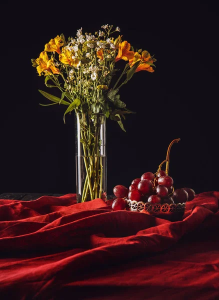 Still life of grapes in metal bowl with flowers in vase on red drapery — Stock Photo