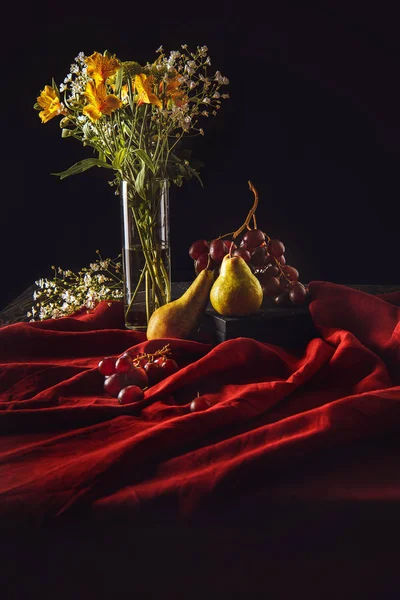 Bodegón con frutas maduras y flores en jarrón sobre tela roja sobre negro - foto de stock