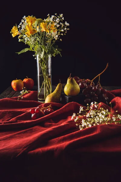 Still life with fruits and flowers in vase on red drapery on black — Stock Photo