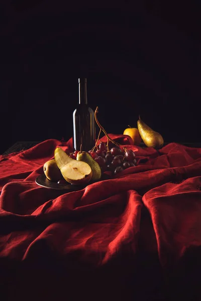 Still life with different fruits and wine bottle on red drapery on black — Stock Photo