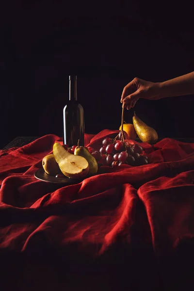 Cropped shot of woman taking grapes from table with wine and pears on black — Stock Photo