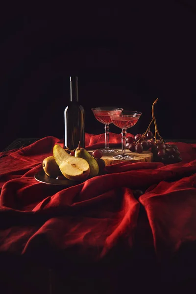 Bodegón con peras, uvas y vino en botella y copas de cristal sobre tela roja sobre negro - foto de stock