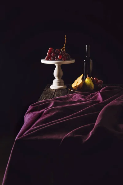 Натюрморт с фруктами и бутылкой вина на бордовых драпировках на черном — стоковое фото
