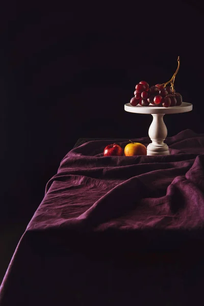 Stand with grapes and apples on table with drapery on black — Stock Photo