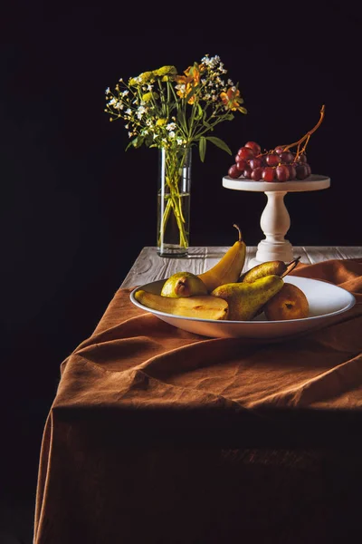 Assiette de poires et stand avec raisins et vase de fleurs sur table sur noir — Photo de stock