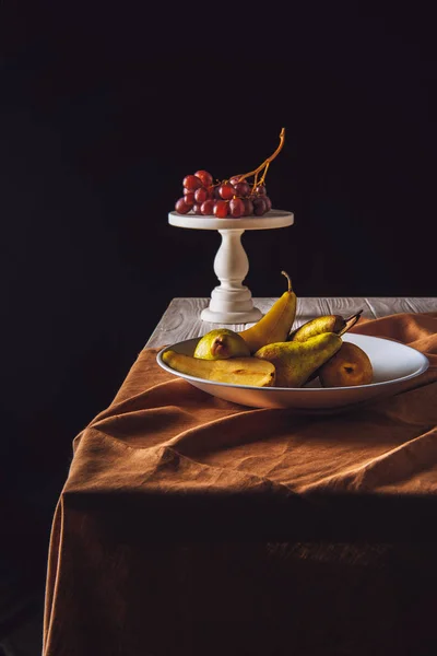 Plato de peras y soporte con uvas sobre mesa sobre negro - foto de stock