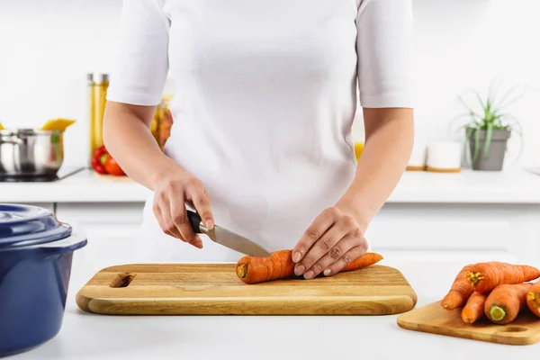 Cropped image of woman cutting carrot on wooden board in light kitchen — Stock Photo