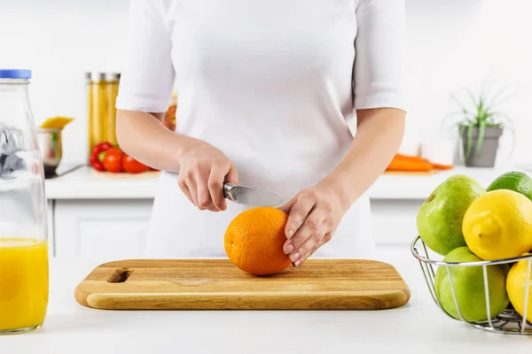 Cropped image of woman cutting orange on wooden board in light kitchen — Stock Photo
