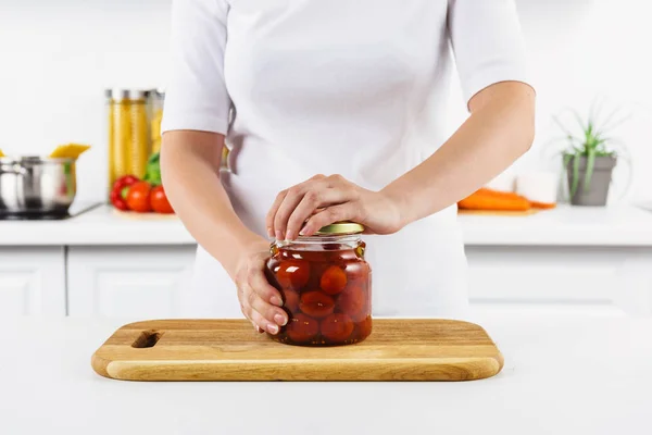 Cropped image of woman opening glass jar with preserved tomatoes in light kitchen — Stock Photo