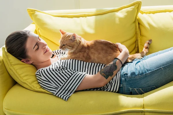 Hermosa sonriente joven acostada en un sofá amarillo con lindo gato rojo — Stock Photo
