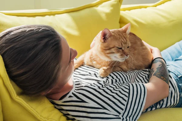 Jovem mulher segurando bonito gato vermelho e deitado no sofá amarelo — Fotografia de Stock